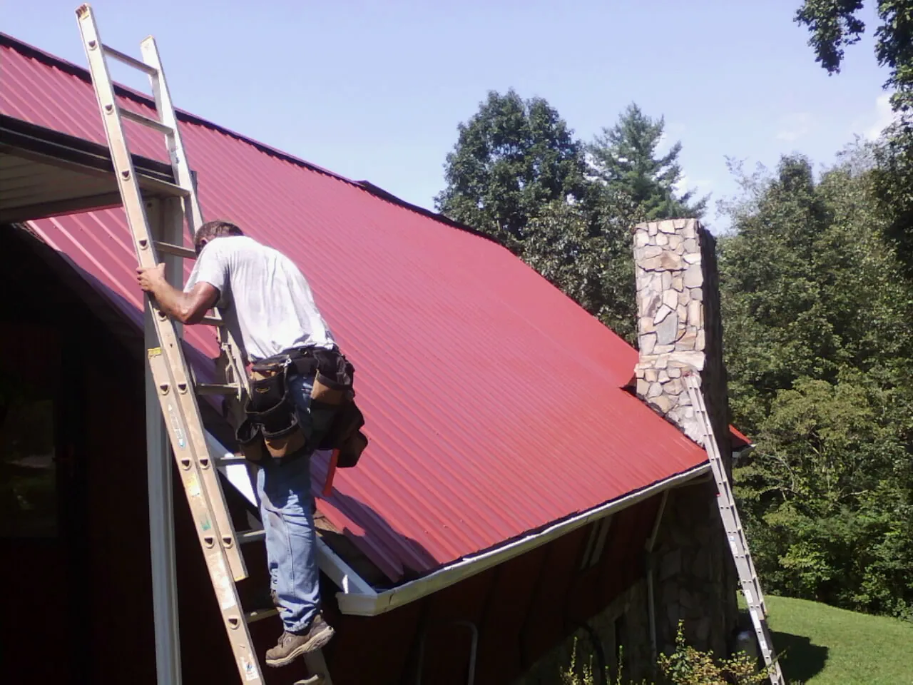 Roofer on a ladder beside a red metal roof and stone chimney — Soffit & Fascia Repair by Roofing Brothers in Constable